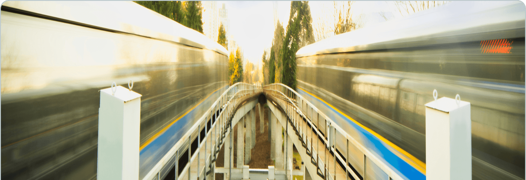 Two blurred trains speeding past a narrow platform with white railings and yellow-blue stripe, trees visible in background.