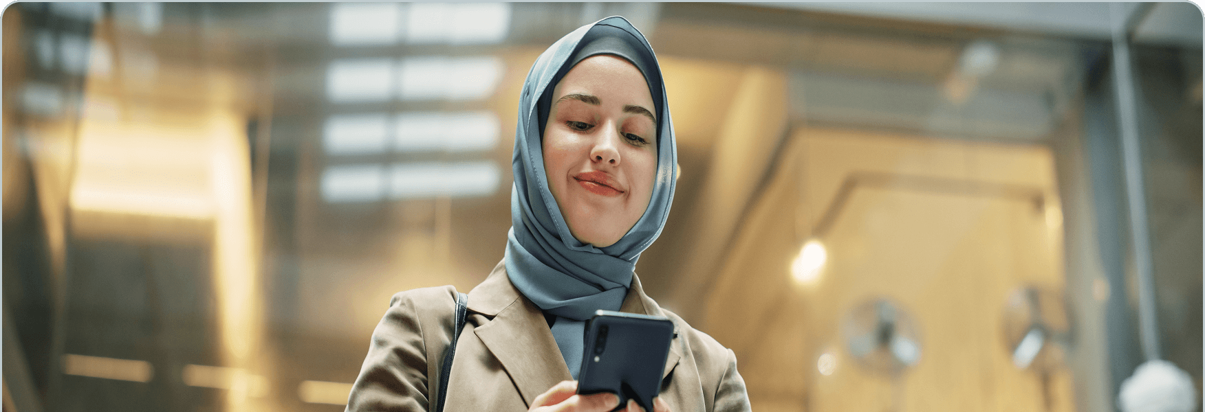 A young woman wearing a blue hijab and beige blazer smiles while looking at her smartphone indoors.